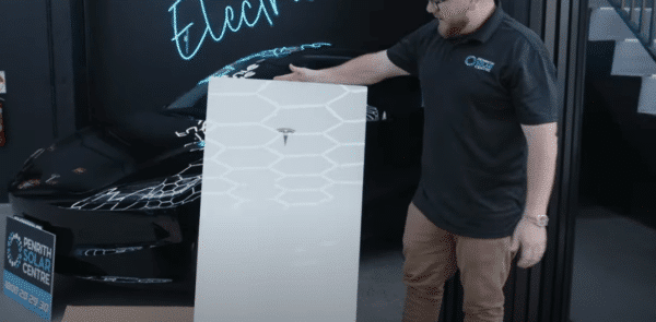 A man stands next to a large Tesla battery in front of a black car, with electric-themed signage in the background.